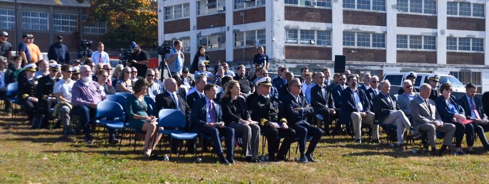Military personnel, public officials, and guests seated outdoors at a military microgrid ribbon-cutting ceremony.