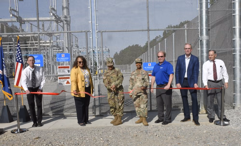 Military personnel and civilian officials hold a red ribbon in front of fenced electrical-substation equipment at a commissioning event.