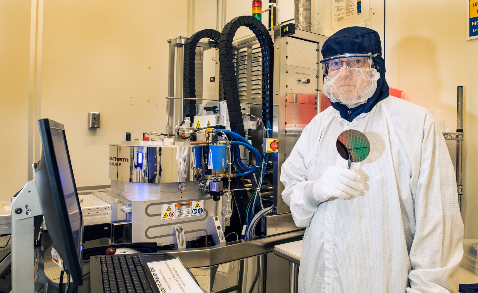 A cleanroom researcher holds a patterned wafer beside a plasma-enhanced chemical vapor deposition tool.